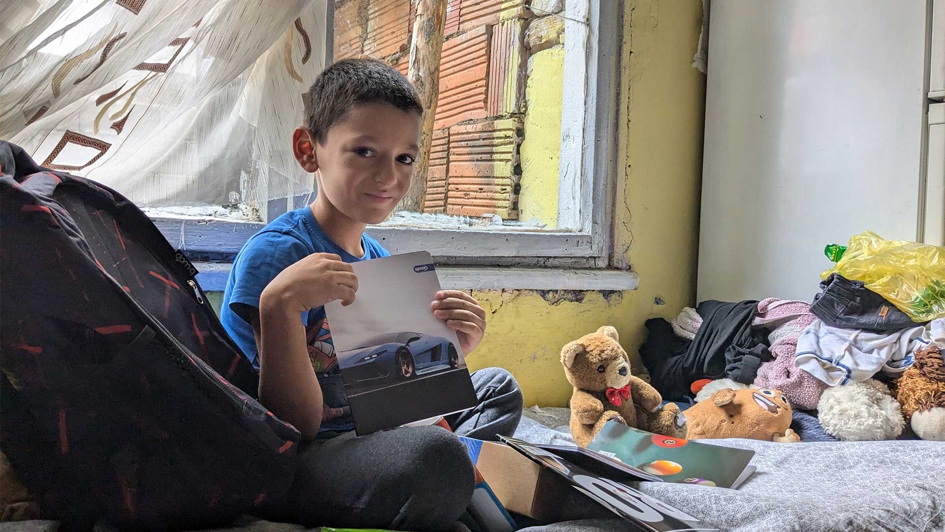 A young boy sits on a bed looking through his new backpack.