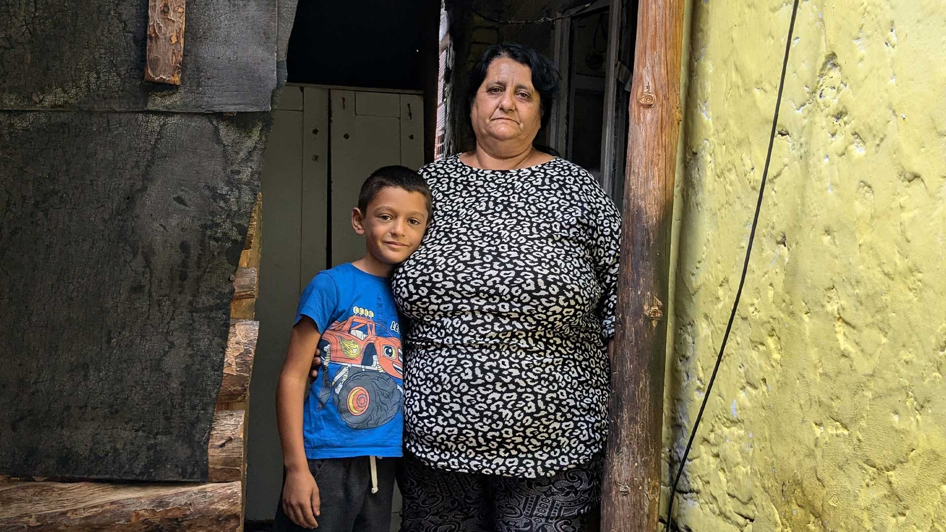 A boy and his grandmother stand outside their small home.