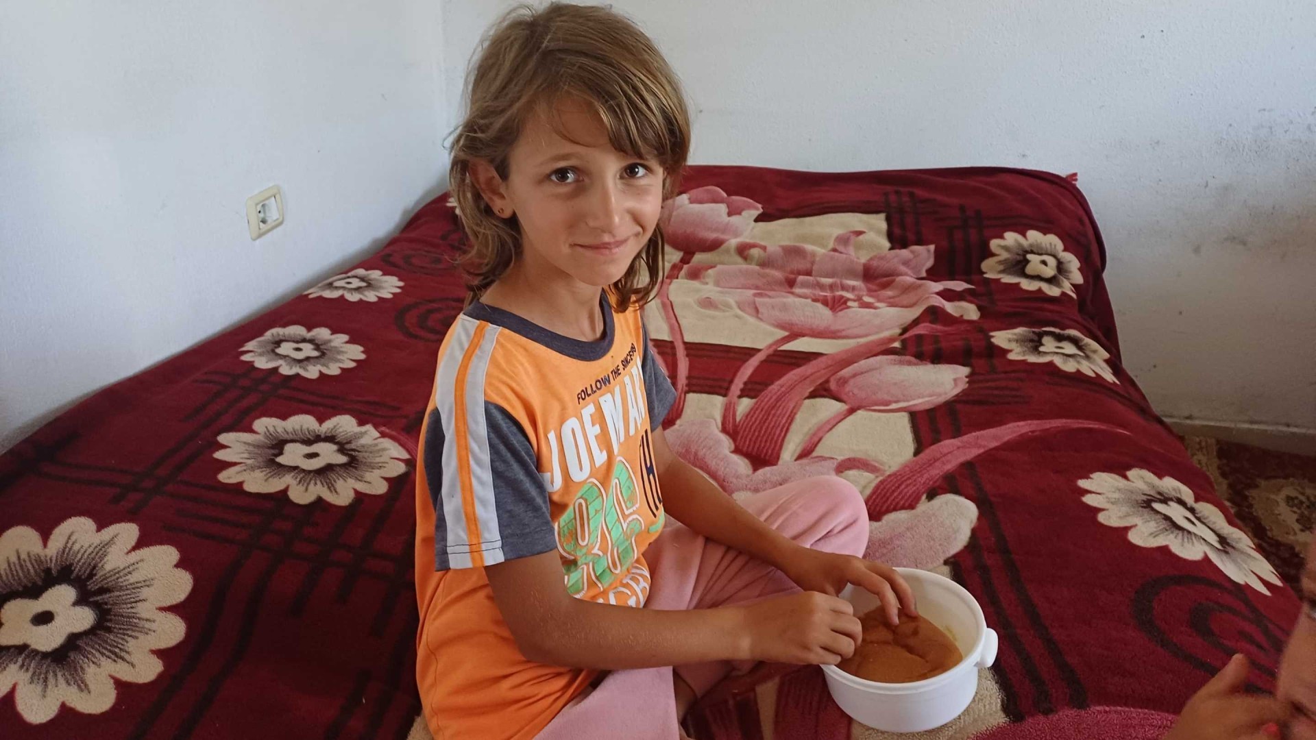 A young girl sits on a bed feeding her brother.