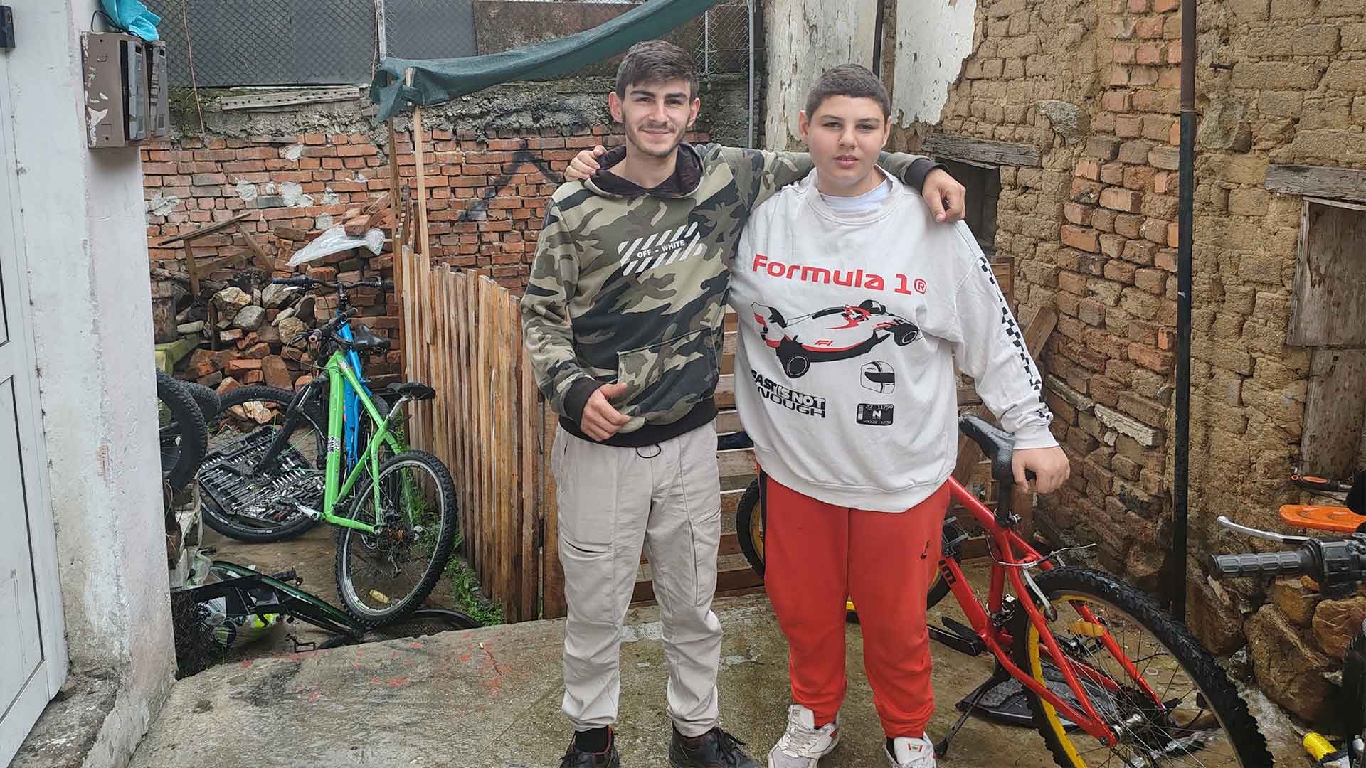 Two brothers stand outside repairing bikes.