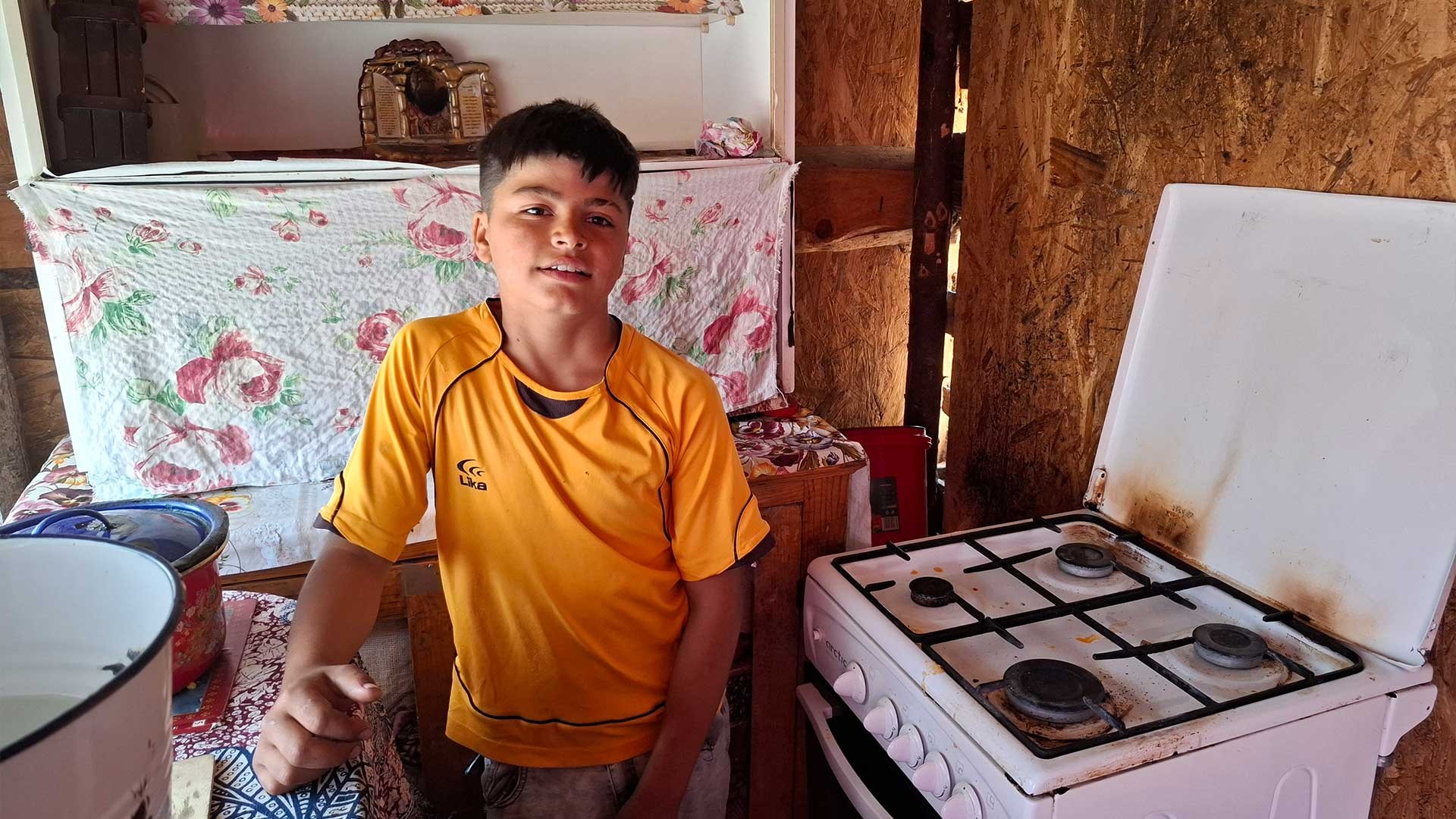 A boy stands in a small kitchen, with holes in the walls.