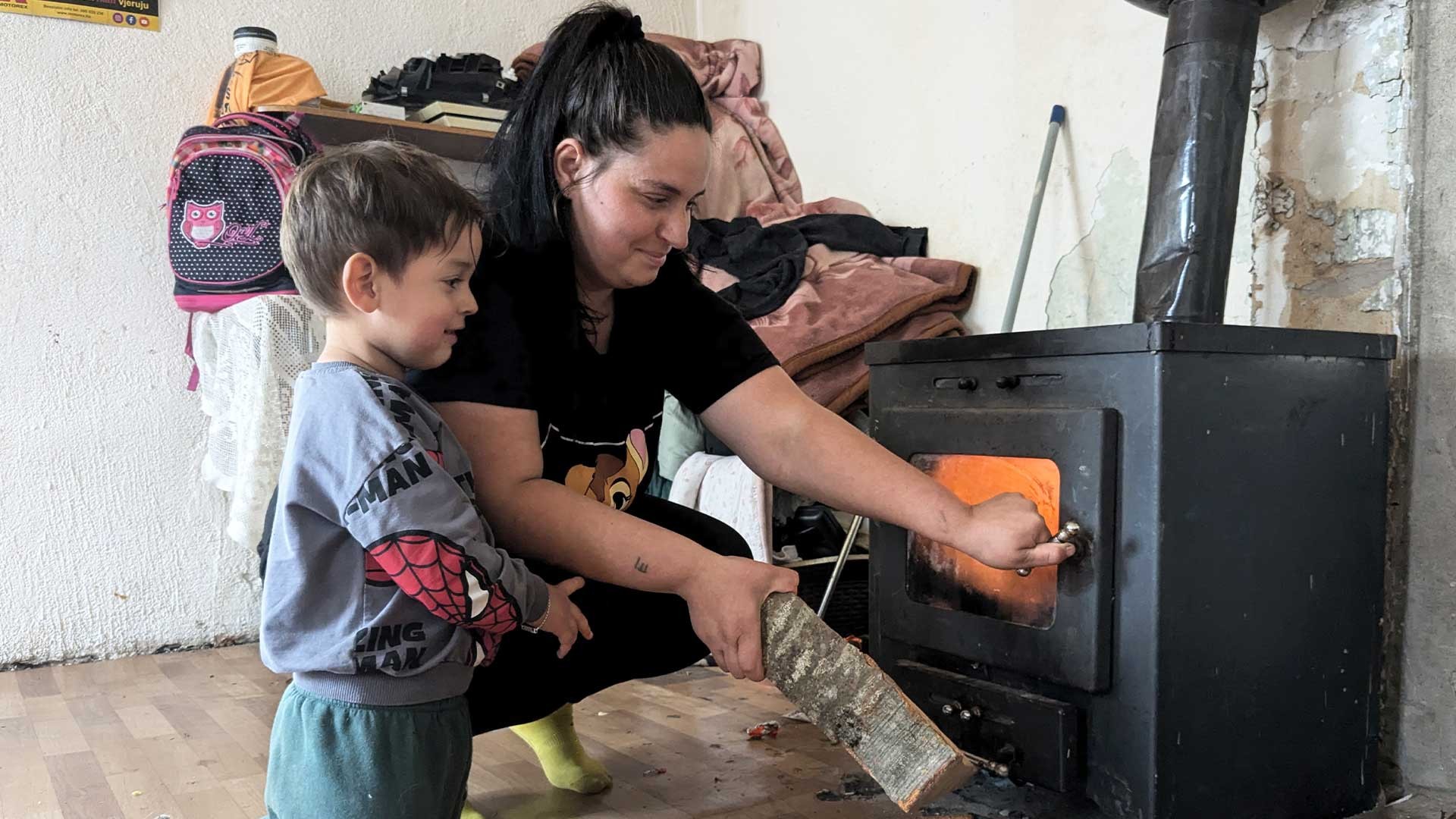 A woman adds wood to a log burner to warm the home.