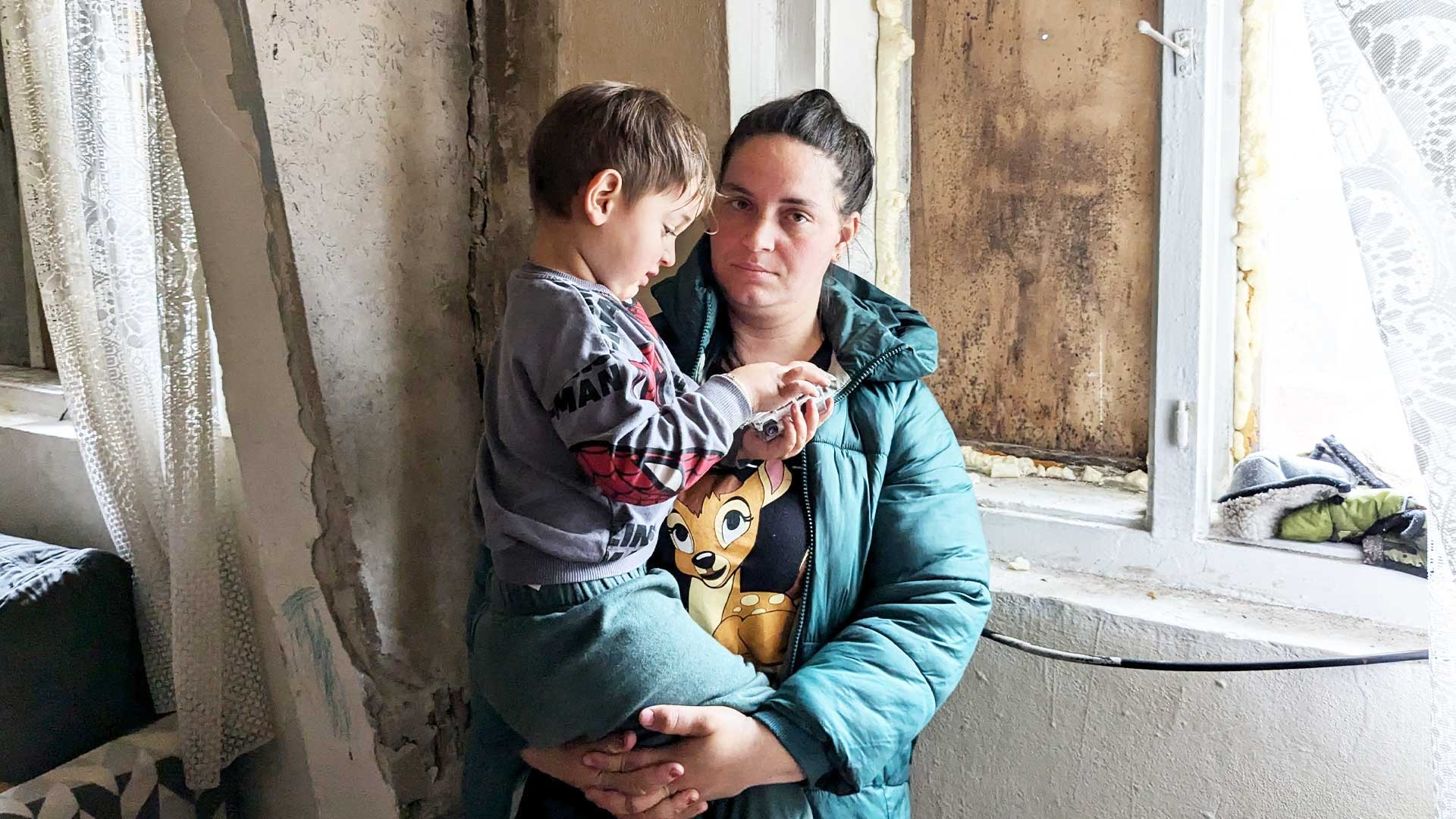 A mother holds her child, standing by a draughty window.